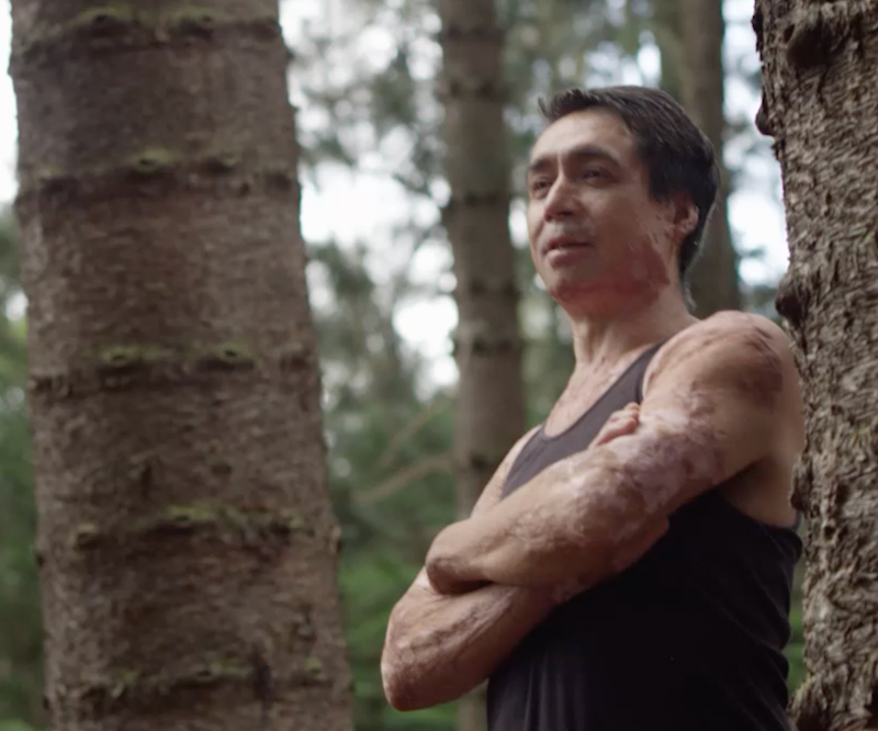 Lance Kaanoi stands in a forest of trees with his arms crossed, looking with determination off into the distance